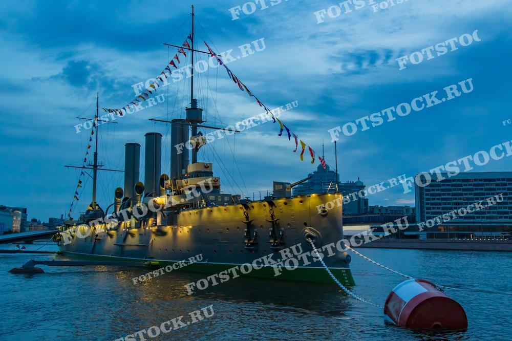 На фотографии изображен крейсер Аврора, освещенный вечерними огнями. Легендарный корабль стоит на Неве, привлекая внимание своим величием и исторической значимостью. Вечернее небо и отражения на воде добавляют снимку атмосферу спокойствия и торжественности. Этот вид подчеркивает важность Авроры как символа революции и музея.